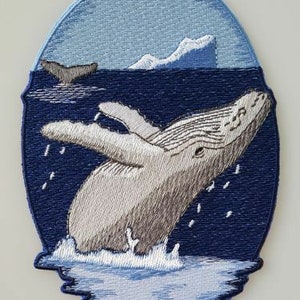 May include: Embroidered patch of a humpback whale breaching out of the water. The whale is white and gray with a blue background. The whale is surrounded by white waves and a blue sky with white clouds.