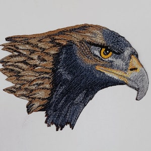 Op de afbeelding: Gestikt patch van een kop van een steenarend met een scherpe snavel en een doordringende gele oog. De veren van de adelaar zijn gedetailleerd in bruin en zwart.