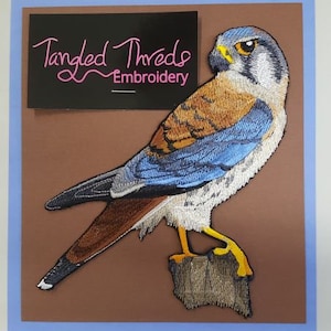 May include: Embroidered patch of a blue and brown American Kestrel bird with yellow feet, perched on a branch. The patch is from Tangled Threads Embroidery.
