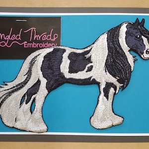 Op de afbeelding: Gestikt patch van een zwart-wit paard met een zilveren manen en staart. Het patch is op een blauwe achtergrond en heeft de tekst "Tangled Threads Embroidery" in de linkerbovenhoek.