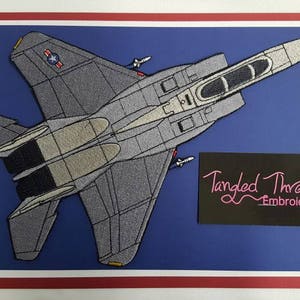 May include: A gray embroidered patch of an F-15 Eagle fighter jet with the text "Tangled Threads Embroidery" on a black background.