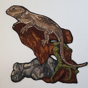Peut inclure: Patch brodé représentant un lézard brun perché sur une branche avec des feuilles vertes. Le lézard a une longue queue et est détaillé avec des écailles.