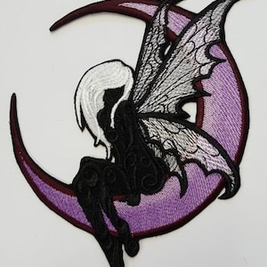 May include: An embroidered patch of a fairy sitting on a purple crescent moon. The fairy has black wings with white accents and a white body with black details.