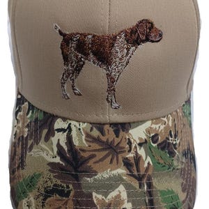 Puede incluir: Una gorra de béisbol de color beige con una visera de camuflaje. La parte delantera de la gorra tiene un puntero alemán de pelo corto bordado en marrón y blanco.