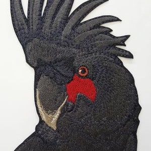 Puede incluir: Parche bordado en negro y rojo de un cacatúa negro con un ojo y un pico rojos.