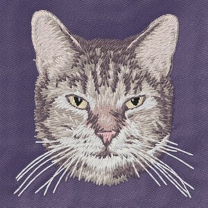 May include: Embroidered cat portrait on a purple background. The cat has gray and brown tabby markings, yellow eyes, and long white whiskers. The embroidery style gives a textured, handmade appearance.