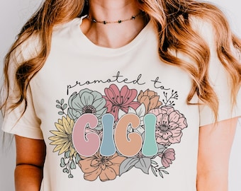 May include: A white t-shirt with the text "promoted to Gigi" in colorful letters surrounded by flowers.