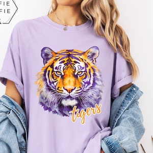 Könnte beinhalten: Ein hellviolettes Kurzarm-T-Shirt mit einer Tigerkopf-Grafik im Aquarellstil in Orange-, Gelb- und Violetttönen. Das Wort "tigers" ist in einer kursiven gelben Schrift unter dem Gesicht des Tigers auf der Vorderseite des T-Shirts aufgedruckt.