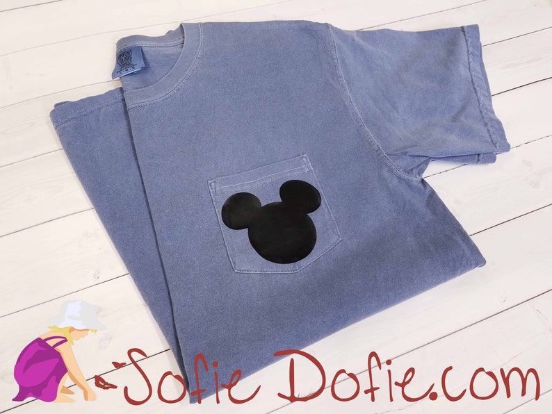 K&ouml;nnte beinhalten: Ein hellblaues T-Shirt mit einer schwarzen Mickey-Maus-Silhouette auf der Tasche.