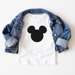 Könnte beinhalten: Ein weißes T-Shirt mit einer schwarzen Mickey Mouse-Silhouette auf der Vorderseite. Das Shirt ist gefaltet und liegt auf einer weißen Oberfläche, darüber liegt eine Jeansjacke.
