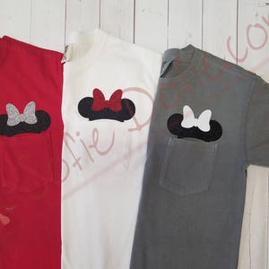 Könnte beinhalten: Drei T-Shirts mit einer Tasche auf der linken Seite. Jede Tasche hat einen andersfarbigen Minnie Mouse Kopf mit Schleife. Die T-Shirts sind rot, weiß und grau.
