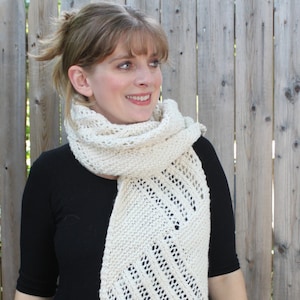 May include: A woman wearing a white knitted scarf with a lacy pattern. The scarf is draped around her neck and hangs down her chest. She is wearing a black shirt and is standing in front of a wooden fence.