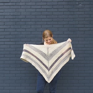 May include: A person is holding a triangular shawl with a beige and purple color scheme. The shawl has a lacy pattern and is made of yarn.
