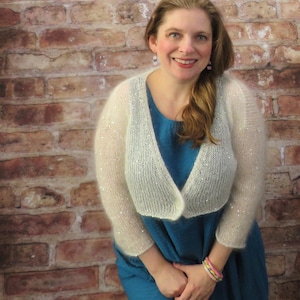 May include: A woman wearing a white, open-front, knitted shrug with sequin accents over a teal dress. The shrug has long sleeves and a V-neck design. The background is a brick wall.
