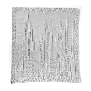 May include: A gray knitted throw blanket with a cityscape design. The blanket is made of a soft, textured yarn and features a variety of different building shapes.
