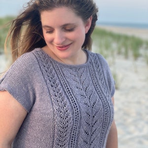 May include: A woman wearing a light purple knitted top with a delicate lace pattern. The top has short sleeves and a round neckline. The woman is standing on a sandy beach.
