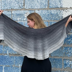 May include: A gray and white ombre knitted shawl with a scalloped edge. The shawl is draped over a person's shoulders.