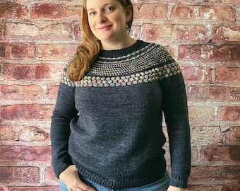 May include: A dark blue sweater with a colourful patterned yoke and long sleeves. The sweater is paired with blue jeans. The background is a brick wall.