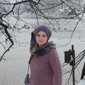 May include: A woman wearing a purple knitted hat and scarf with a matching purple sweater. The hat has a long scarf with a tassel. The woman is standing in front of a snowy background.