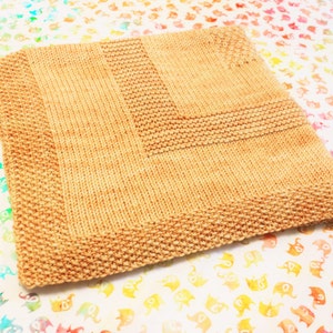 May include: A tan knitted baby blanket with a textured pattern. The blanket is folded in half and lying on a white surface with a colorful pattern.