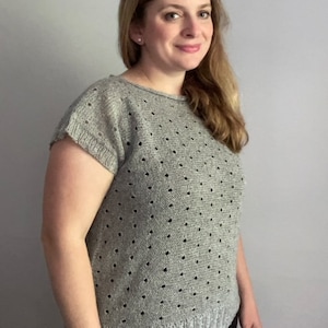 Puede incluir: Una blusa gris de manga corta de punto con un patrón de puntos. La blusa tiene un escote redondo y un ajuste ligeramente holgado.