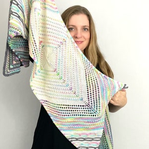 May include: A white crocheted shawl with colorful stripes and a lacy pattern. The shawl is held up by a person, partially obscuring their face.