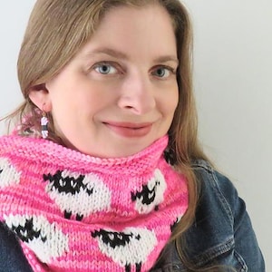 May include: A pink and white knitted cowl featuring a sheep pattern. The cowl is made of soft yarn and is designed to be worn around the neck. The sheep have black faces and legs. The background is a light pink.