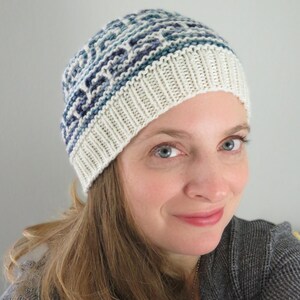 May include: A white and blue knitted hat with a geometric pattern. The hat is worn by a person with long brown hair.