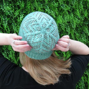 May include: A light teal knitted hat with a patterned design. The hat is held up by two hands, with a green leafy background. The hat has a brim and a detailed stitch pattern with brown accents.