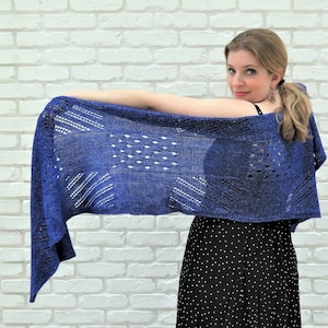 May include: A blue knitted shawl with a geometric openwork design. The shawl is a deep shade of blue and features a variety of patterns, including rows of small holes and diagonal lines. The shawl is being held up, showcasing its length and texture.