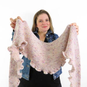 Könnte beinhalten: Ein dreieckiges Stricktuch in hellem Cremeweiß mit bunten Sprenkeln. Das Tuch hat eine kraus rechts gestrickte Textur und einen gerüschten Rand entlang der unteren Kante, was an beiden Seiten des Kleidungsstücks einen wellenförmigen Abschluss erzeugt.