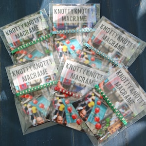 May include: Several clear plastic packages of macrame bracelets are displayed. Each package has a card with the text "KNOTTY KNOTTY MACRAME." The bracelets are in various colors, including green, red, and blue. The background is a blue surface.