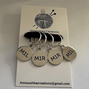 May include: A set of four silver-colored stitch markers with the text "M1L" and "M1R" engraved on them. The markers are attached to a black fuzzy band and a white card with the logo "Annoushka Creations" and the email address annoushkacreations@gmail.com.