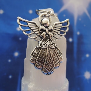 May include: A silver angel charm with intricate details and a bow on its chest. The charm is hanging from a key ring and is resting on a white crystal.