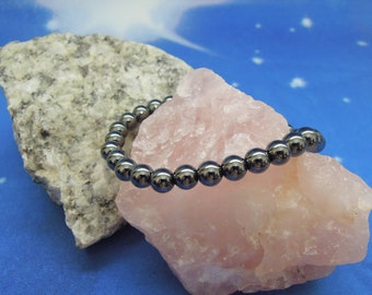 May include: A black hematite beaded bracelet on a pink and gray rock background.