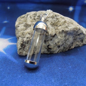 May include: A clear glass vial pendant with silver-colored metal caps on each end, suspended on a delicate chain. The vial rests on a gray and white stone against a blue background with a starburst design.