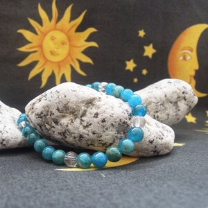 May include: A bracelet with turquoise and clear crystal beads. The bracelet is displayed on a textured stone. The background is a celestial-themed fabric with a sun, moon, and stars.