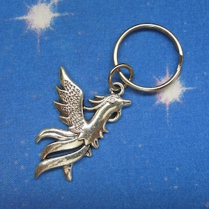 Puede incluir: Llavero de plata con un diseño de fénix detallado. El fénix está mirando hacia la derecha con las alas extendidas. El llavero está unido a un anillo de plata.