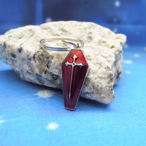Puede incluir: Un llavero rojo en forma de ataúd con un diseño de cruz plateada. El llavero es de metal y tiene un llavero plateado. El fondo es una tela azul con una roca blanca.