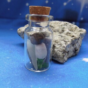 May include: A small, clear glass bottle with a cork stopper containing several smooth, polished stones in shades of white, light pink, dark brown, and green. The bottle is cylindrical with a narrow neck and a flat base, holding the assorted pebbles.