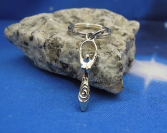 May include: A silver-toned metal keychain featuring a stylized female figure with arms raised in a circular shape above the head and a spiral design at the base. The charm is attached to a standard circular metal split ring for keys.