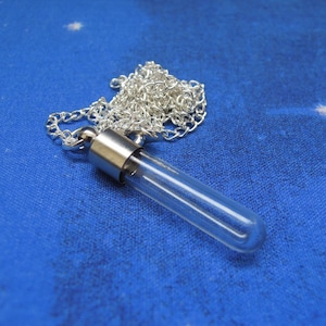 May include: A clear glass vial pendant necklace with a silver-colored metal cap and chain. The vial is cylindrical and empty. The necklace rests on a blue fabric background.