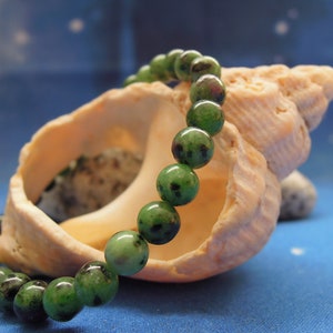 Puede incluir: Un brazalete de piedras preciosas verdes con motas verde oscuro, descansando sobre una concha. El brazalete está hecho de cuentas redondas y se muestra sobre un fondo azul.