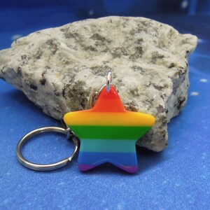Könnte beinhalten: Ein regenbogenfarbener sternförmiger Schlüsselanhänger mit einem silbernen Schlüsselring. Der Stern hat die Farben des Regenbogens, mit Rot oben, das in Gelb, Grün, Blau und Lila übergeht. Der Schlüsselanhänger befindet sich vor einem blauen Hintergrund.