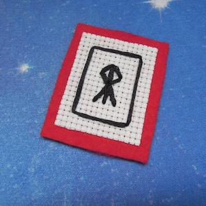 May include: A rectangular patch featuring a white Aida cloth center with a black embroidered rectangular border and a central black stitched symbol, all framed by a solid red felt border. The design is flat and has a visible grid texture.
