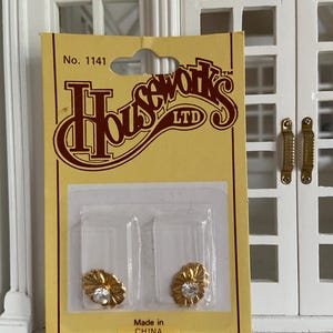May include: A pair of gold-toned, flower-shaped decorative knobs with a central clear crystal, packaged on a yellow Houseworks LTD No. 1141 card. The packaging includes the text Made in CHINA and the address 2388, PLEASANTDALE ROAD, ATLANTA, GEORGIA 30340.
