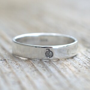 Puede incluir: Un anillo de banda de plata con un s&iacute;mbolo simple grabado en la parte superior. El s&iacute;mbolo es un c&iacute;rculo con una l&iacute;nea que lo atraviesa, y un c&iacute;rculo m&aacute;s peque&ntilde;o dentro del c&iacute;rculo m&aacute;s grande.