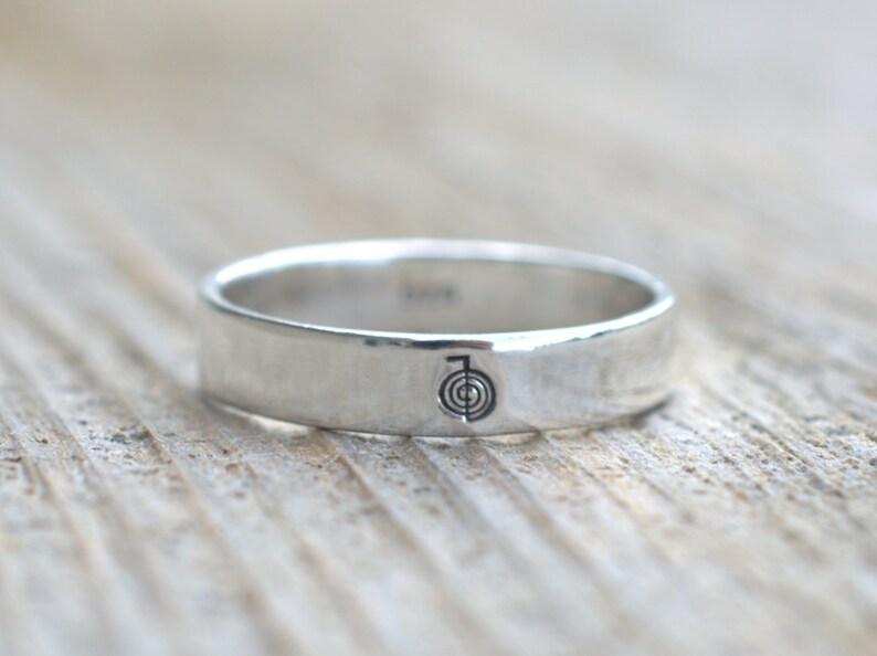 Puede incluir: Un anillo de banda de plata con un s&iacute;mbolo simple grabado en la parte superior. El s&iacute;mbolo es un c&iacute;rculo con una l&iacute;nea que lo atraviesa, y un c&iacute;rculo m&aacute;s peque&ntilde;o dentro del c&iacute;rculo m&aacute;s grande.
