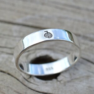 Puede incluir: Un anillo de banda de plata con un s&iacute;mbolo grabado simple en la parte superior. El anillo est&aacute; sobre una superficie de madera.