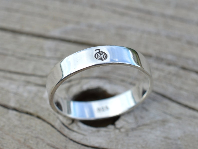 Puede incluir: Un anillo de banda de plata con un s&iacute;mbolo grabado simple en la parte superior. El anillo est&aacute; sobre una superficie de madera.
